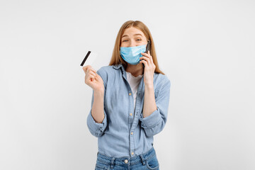 Happy young woman in a medical mask on her face, showing a plastic credit card and talking on a mobile phone, on a white background