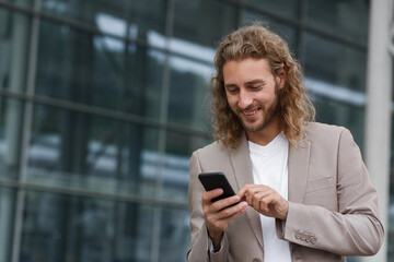 Portrait of handsome curly businessman in casual wear holding smartphone and smiling.Successful manager using mobile phone apps,texting message,browsing internet,looking at phone near business center.