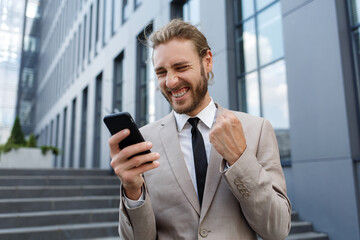 Excited and happy businessman winner is holding a smartphone in euphoria from victory.Portrait of a curly-haired young businessman manager in a white shirt on the background of a business center.