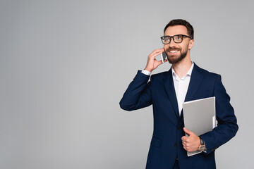 bearded businessman in blue blazer and eyeglasses looking away while talking on smartphone isolated on grey