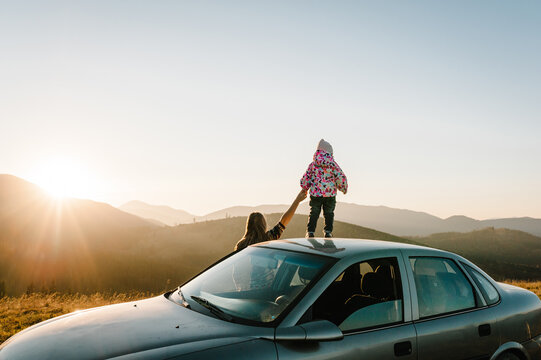 Mother and child in mountains on the sunset near car. Mom and daughter - tourist girls. Toward adventure! Family enjoying road trip on autumn vacation. Holiday and travel concept. Back view.