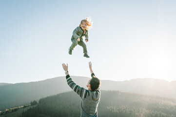 Dad playing with daughter and throws her high in the sky in mountains at the sunset time. Concept of friendly family. Portrait Dad and child enjoying autumn vacation. A place for text, advertising.