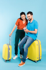 young man in polo t-shirt using smartphone while sitting on suitcase near asian woman in red blouse on blue