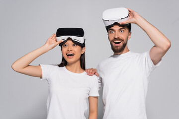 excited interracial couple in white t-shirts looking away with open mouths while touching vr headsets on grey