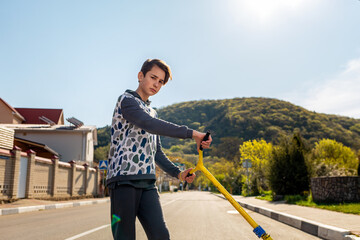 A confident teenage boy with an arrogant look and a scooter in his hand.In the background,a street and road on a Sunny day,a view of the mountain.Copy space.Concept of sports and adolescent activities