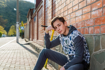 A teenage boy sits on the sidewalk next to a scooter and poses leaning on his arm. Brick wall and street in the background. The concept of adolescence and youth activity