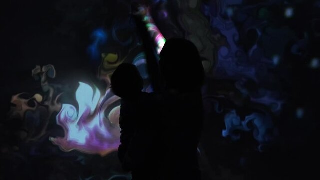 Girl with kid plays with an interactive video installation. New art form, generative graphics. Silhouette of girl with kid draws multi-colored paints interactive installation.