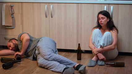 Helpless beaten woman suffering sitting on the floor near drunk man. Alcoholic aggressive husband abusing injuring terrified, afraid, beaten and panicked wife living in terror and pressure.