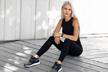 Beautiful blonde female in black sportswear resting after workout exercises outdoors. Fitness girl takes a rest after running in the morning sitting agaisnt white wall outside.