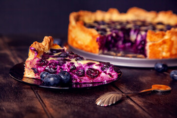 Rustic shortbread pie with blueberries in sour cream filling on wooden background with piece cut off on saucer and spoon