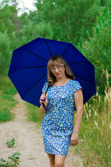A young woman in glasses and a blue dress holds a blue umbrella.