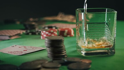 Whiskey Or Rum Is Slowly Poured Into a Glass, And Then Ice Cubes Are Thrown. Casino Poker Chips, Coins And Deck Of Playing Cards Stacked In Multiple Piles On Table. Gambling fortune. Slow motion - Powered by Adobe