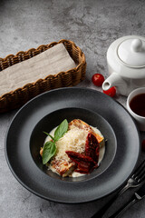 A big piece of italian lasagna in a restaurant, black plate, grey background