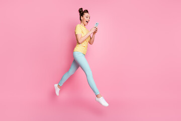 Full size photo of funny pretty lady two buns hold telephone read blog post comments walk street jump high up wear casual striped t-shirt pants shoes isolated pink color background