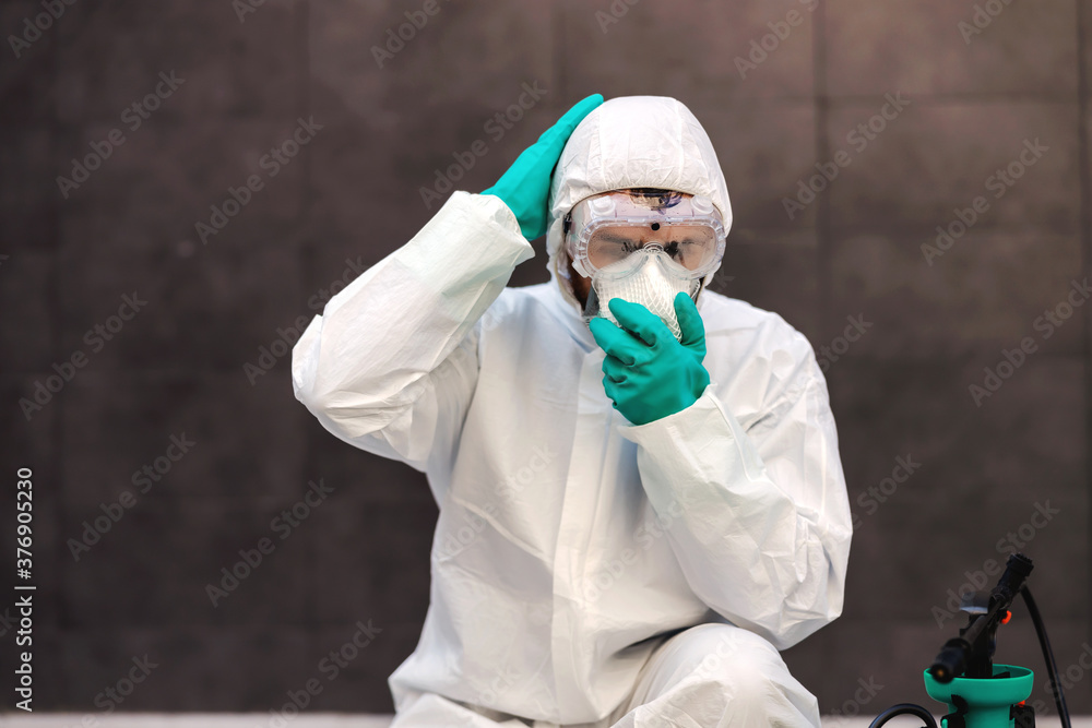 Wall mural Man in sterile protective uniform with rubber gloves crouching outdoors and putting protective mask on face. Next to him is sprayer with disinfectant. Prevention form spreading corona virus concept.