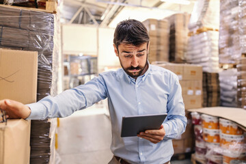 Amazed young attractive bearded supervisor standing in warehouse and looking at tablet.