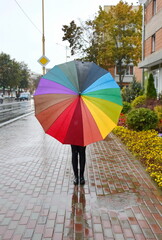 A girl with a rainbow-coloured umbrella, standing outside in rainy weather