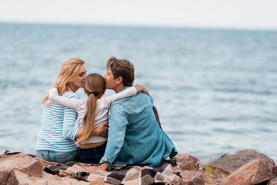 Back view of parents embracing and kissing daughter on plaid near sea