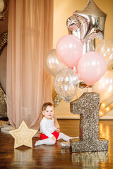First year's birthday. A happy little girl in a white dress and reds shirts sits against a nice background of garlands and pink balloons, celebrating her first birthday. Decoration of the birthday.