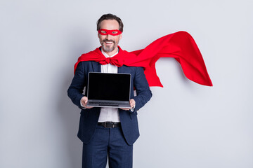 Photo of confident cheerful aged mature business guy super hero costume hold laptop presenting new device model sale price manager wear suit red face mask cloak isolated grey background