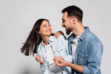 stylish interracial couple in denim shirts holding hands while looking at each other on grey
