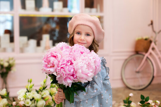 Closeup portrait of happy blonde little girl with long curly hair and in a beret with bouquet of pink peonies. Childhood concept. Celebration. cute child with flowers for mothers day. Celebration.	
