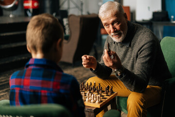 Obraz premium Mature adult bearded gray-haired grandfather explaining rules of chess game to grandchild at home in cozy living room with an authentic aristocratic interior.