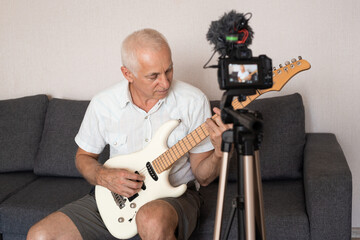 Senior man recording music video blog, home lesson or song, playing guitar or making broadcast internet tutorial while sitting in sofa at home. Concept of hobby, music, art and creation.