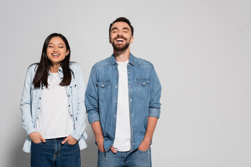 young interracial couple in denim clothes holding hands in pockets while standing with closed eyes on grey