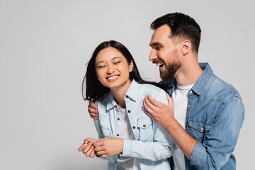excited man touching shoulders of asian girlfriend in denim shirt on grey