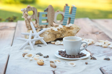 Obraz na płótnie Canvas White cup of espresso coffee on wooden table with sand, shells, starfish and rocks.