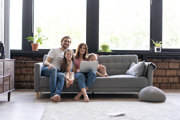 Happy parents hug kids lying on cozy sofa watching cartoons on laptop, young family spend time with children relaxing at home, mom and dad cuddle with daughters enjoying funny video at computer