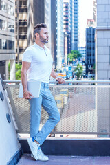 Young Serious European Businessman with beard, little gray hair, wearing white shirt, light blue pants, white sneakers, holding laptop computer, standing by railing on balcony in New York City..