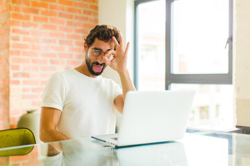young bearded man with a laptop smiling happily with funny face, joking and looking through...