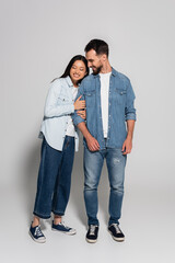 full length view of young asian woman with closed eyes leaning on stylish boyfriend on grey