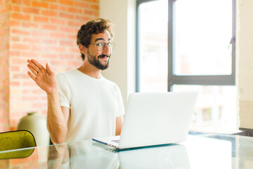 young bearded man with a laptop smiling happily and cheerfully, waving hand, welcoming and greeting you, or saying goodbye