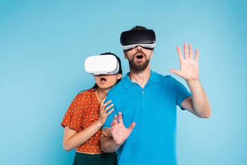 scared woman in red blouse touching excited man in polo t-shirt while using vr headsets together isolated on blue