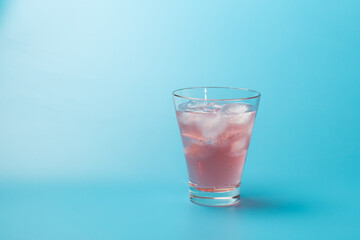 Glass of refreshing pink cocktail or iced tea with ice cubes on blue background with copy space.