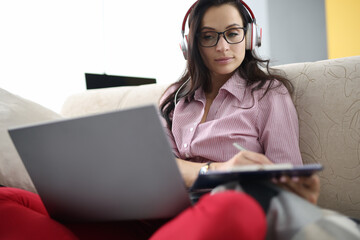 Woman in headphones with clipboard in her hands sits on couch with laptop. Employee transition to...