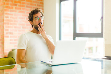 young bearded man with a laptop yawning lazily early in the morning, waking and looking sleepy,...