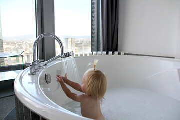 little kid, baby bathes in a large round bath, jacuzzi in a bright room by the window, puts his hands to the water, hygiene concept, luxury body care from childhood - obrazy, fototapety, plakaty