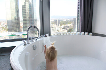 little kid, baby bathes in a large round bath, jacuzzi in a bright room by the window, puts his hands to the water, hygiene concept, luxury body care from childhood - obrazy, fototapety, plakaty