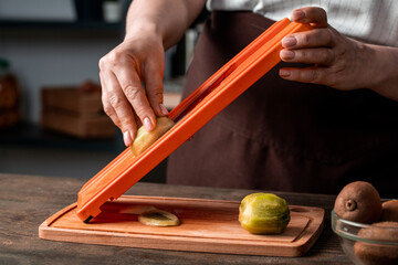 Close-up of unrecognizable woman cutting kiwi on slicer above wooden board in kitchen