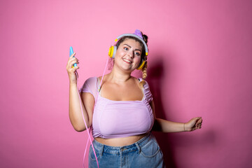 Young plus size woman on pink background dancing holding smartphone - Chubby woman listening steaming music isolated studio shot