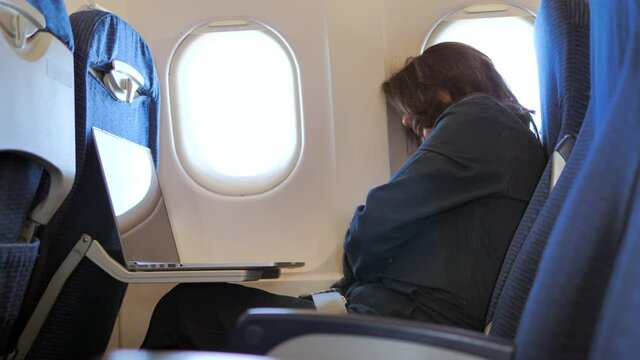 Woman sleeping in an airplane beside window. Tired passenger relaxes in a flying aircraft with Laptop.