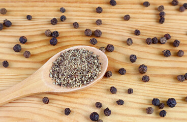 Ground black pepper on wooden spoon with black peppercorns scattered on wooden background