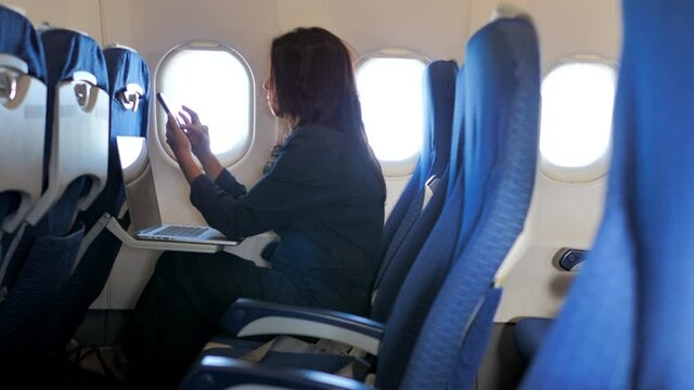 Asian woman sitting in airplane cabin and chatting online on smartphone while checking email on laptop computer while flying at plane.