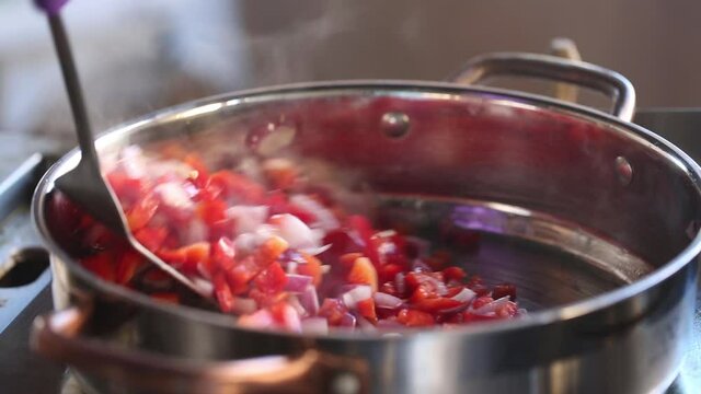 The chef mixes vegetables, onions and tomatoes in a saucepan. Steam rises from the pan, close up