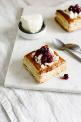 Puff pastry squares with ricotta and cherry sauce on white marble tray. Baked small square cakes topped with cheese and berry sauce