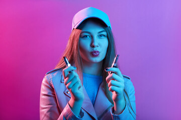 Young beautiful female wearing casual clothing and hat, woman holding two pomades in hands, choosing pomade for makeup, girl keeps lips rounded, poses isolated over pink neon wall.